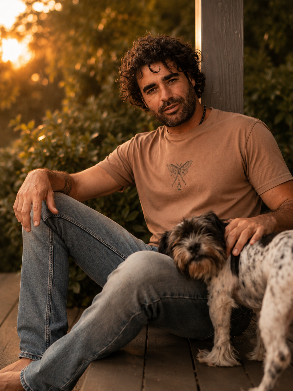 Renan wearing the Son of Nature Noches del Fuego luna moth tee in clay, charcoal luna-moth line art on the chest, golden-hour Kauai deck with a dog