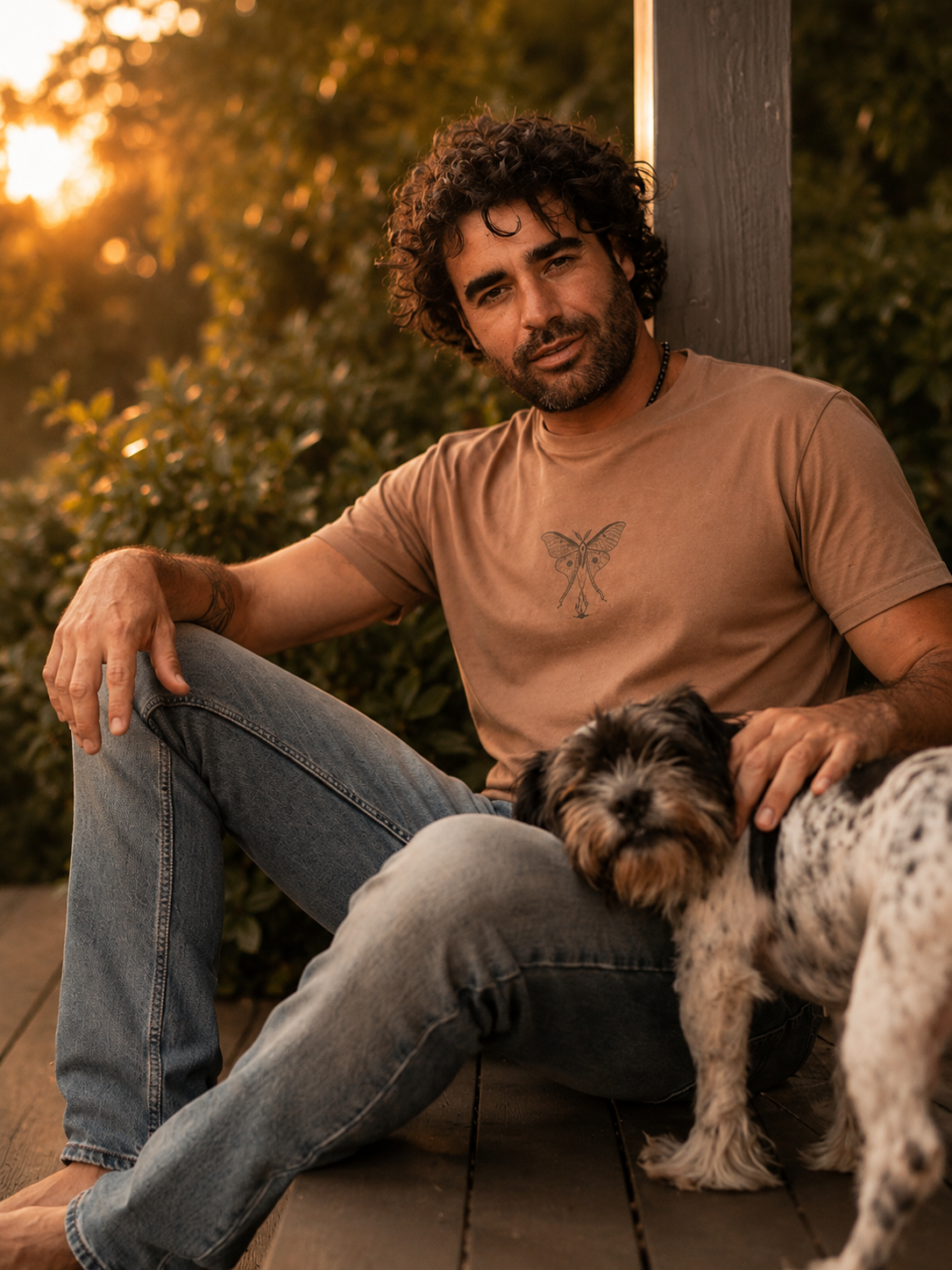 Renan wearing the Son of Nature Noches del Fuego luna moth tee in clay, charcoal luna-moth line art on the chest, golden-hour Kauai deck with a dog
