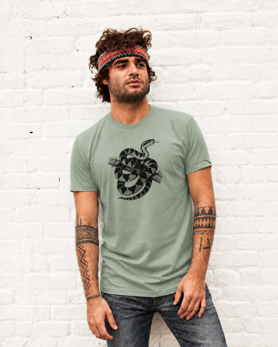 Renan wearing the Son of Nature Surucucu bushmaster tee in bay, charcoal bushmaster coiled on a branch on the chest, against a white brick wall